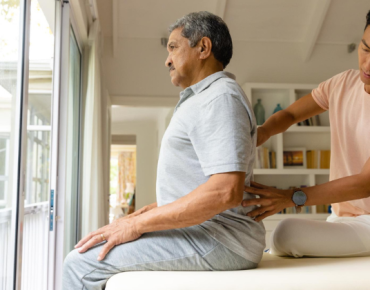 Physiotherapist performing back therapy on an elderly man during a home physiotherapy session in a bright, comfortable living space.