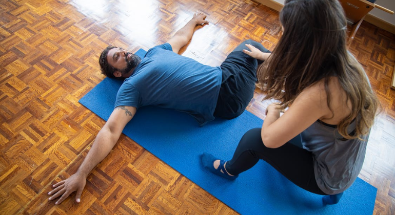 Physiotherapist guiding a man through lower back stretching exercises during a home physiotherapy session on a yoga mat in a wooden-floored room.