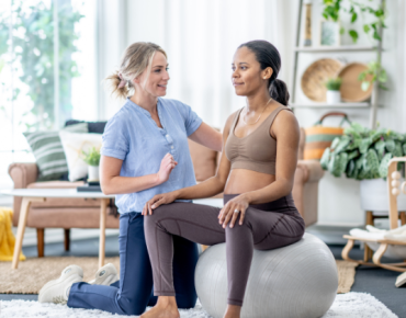 Physiotherapist guiding a pregnant woman through stability exercises using an exercise ball during a home physiotherapy session in a cozy, well-lit living space.