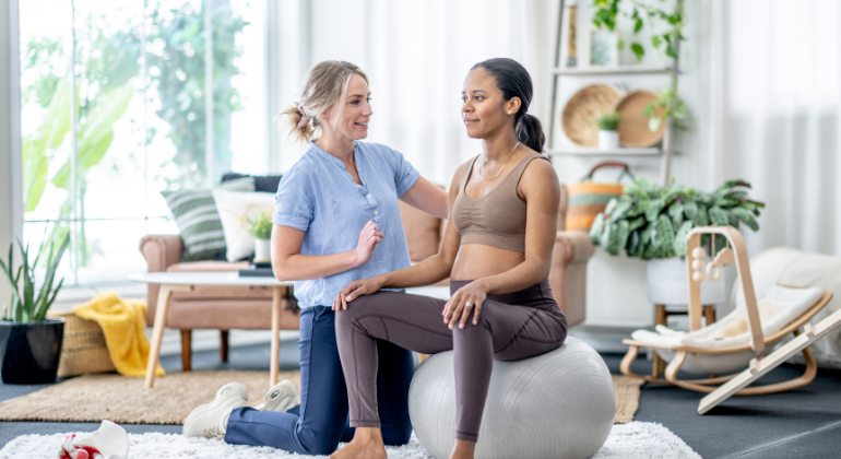 Physiotherapist guiding a pregnant woman through stability exercises using an exercise ball during a home physiotherapy session in a cozy, well-lit living space.