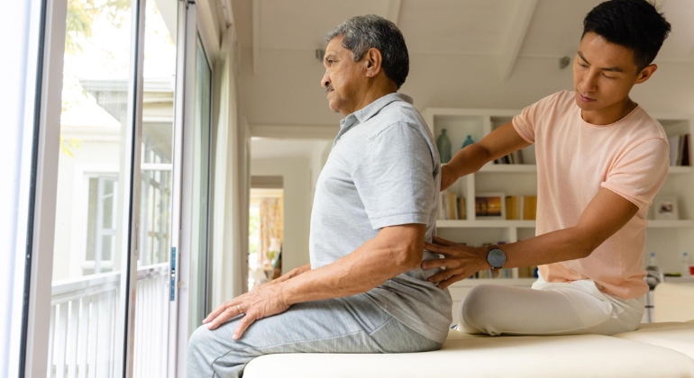 Physiotherapist performing back therapy on an elderly man during a home physiotherapy session in a bright, comfortable living space.