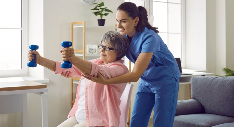 Physiotherapist assisting an elderly woman with arm strengthening exercises using dumbbells during a home physiotherapy session in a bright and comfortable living room.