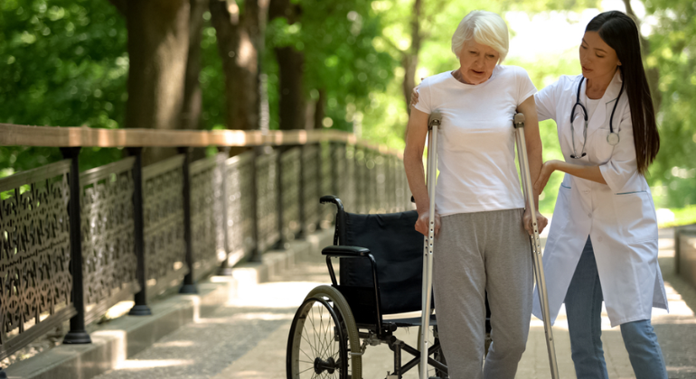 Healthcare professional assisting an elderly woman with crutches during a rehabilitation session outdoors, with a wheelchair nearby for support.