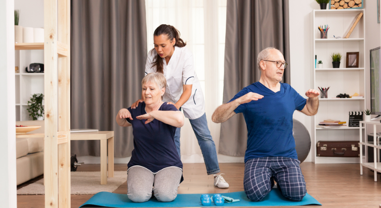 Physiotherapist guiding an elderly couple during a home exercise session, with the couple performing stretches on yoga mats in a well-lit living room.