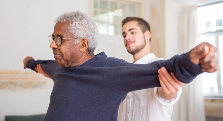 Physiotherapist assisting an elderly man with shoulder and arm mobility exercises during a home physiotherapy session in a bright, relaxed environment.