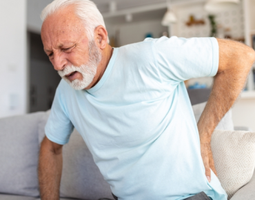 An elderly man sitting on a sofa holding his lower back in pain. He appears to be experiencing discomfort, indicating possible back or joint issues.