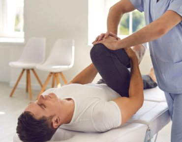 A physiotherapist performing a knee therapy session on a male patient lying on a treatment table in a clinic setting. The therapist is gently bending the patient’s leg to improve mobility.