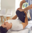 A physiotherapist performing a knee therapy session on a male patient lying on a treatment table in a clinic setting. The therapist is gently bending the patient’s leg to improve mobility.