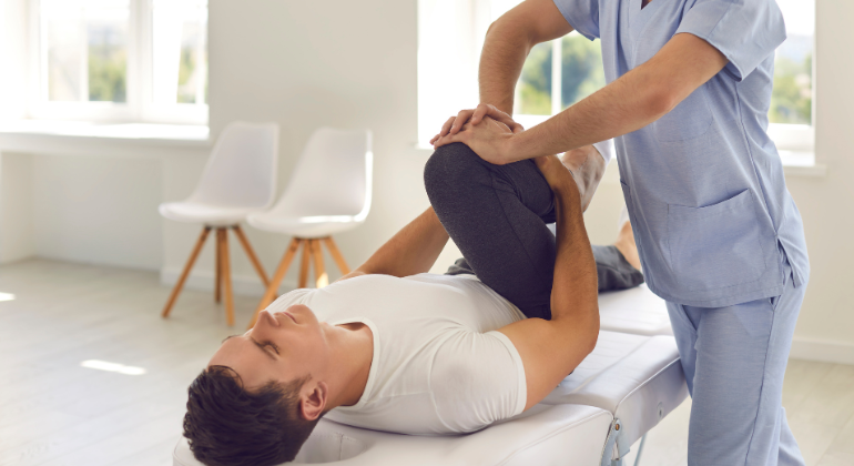 A physiotherapist performing a knee therapy session on a male patient lying on a treatment table in a clinic setting. The therapist is gently bending the patient’s leg to improve mobility.