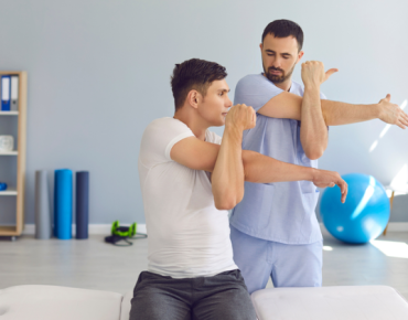 A physiotherapist guiding a male patient through shoulder stretching exercises. The patient is sitting on a treatment table, extending his arm across his chest in a clinic setting.