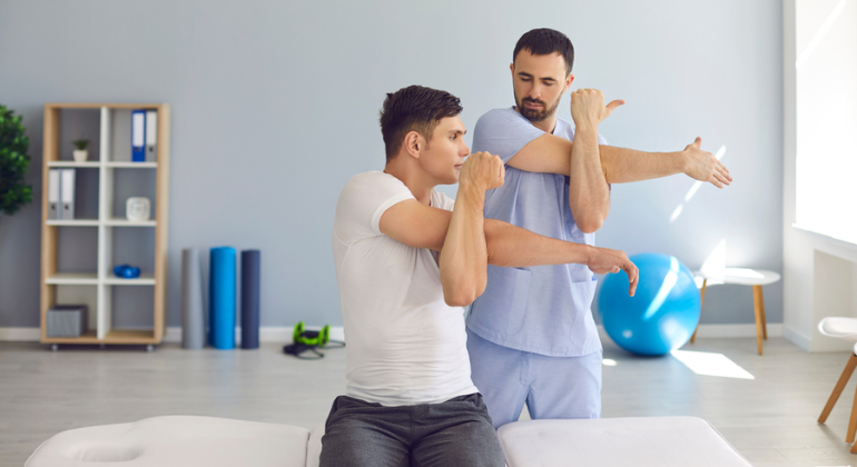 A physiotherapist guiding a male patient through shoulder stretching exercises. The patient is sitting on a treatment table, extending his arm across his chest in a clinic setting.