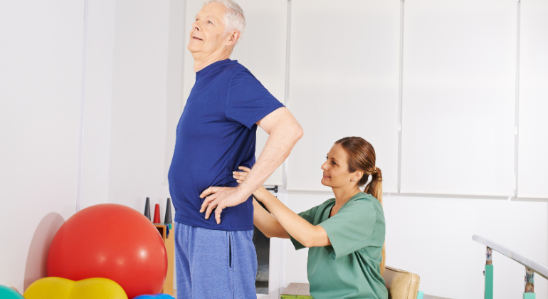 A physiotherapist assisting an elderly man with back rehabilitation exercises. The man is standing with his hands on his hips while the therapist supports his lower back during the session.