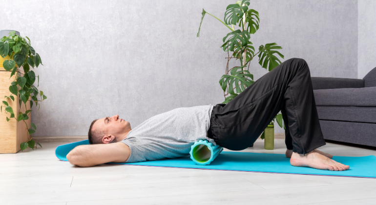 A man lying on a yoga mat using a foam roller under his lower back for self-massage and muscle relaxation. The exercise is performed in a home setting with plants in the background.