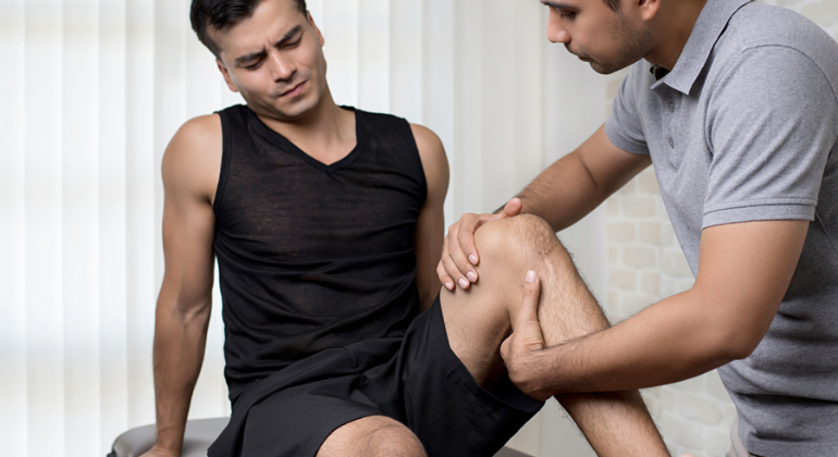 A physiotherapist examining a male patient's knee during a therapy session. The patient, sitting on a treatment table, appears to be in discomfort as the therapist assesses the joint for mobility and pain.