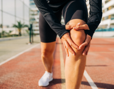 A person holding their knee in discomfort while standing on a sports track, indicating knee pain or injury. The individual is dressed in athletic wear, suggesting activity-related strain.