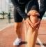 A person holding their knee in discomfort while standing on a sports track, indicating knee pain or injury. The individual is dressed in athletic wear, suggesting activity-related strain.