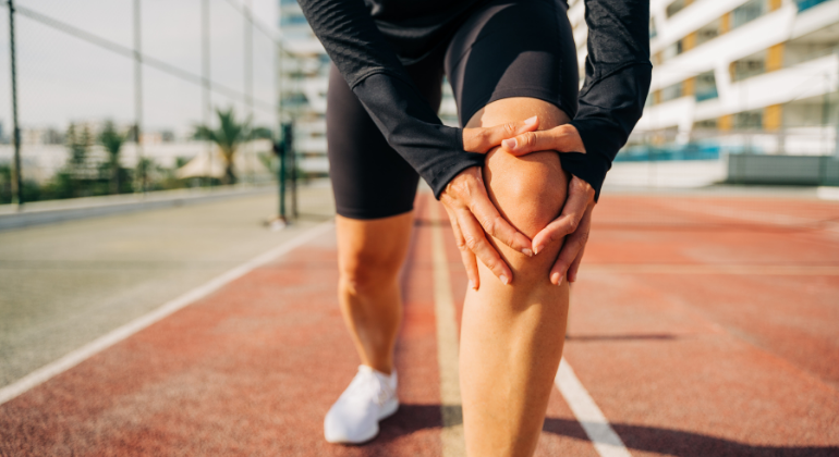 A person holding their knee in discomfort while standing on a sports track, indicating knee pain or injury. The individual is dressed in athletic wear, suggesting activity-related strain.