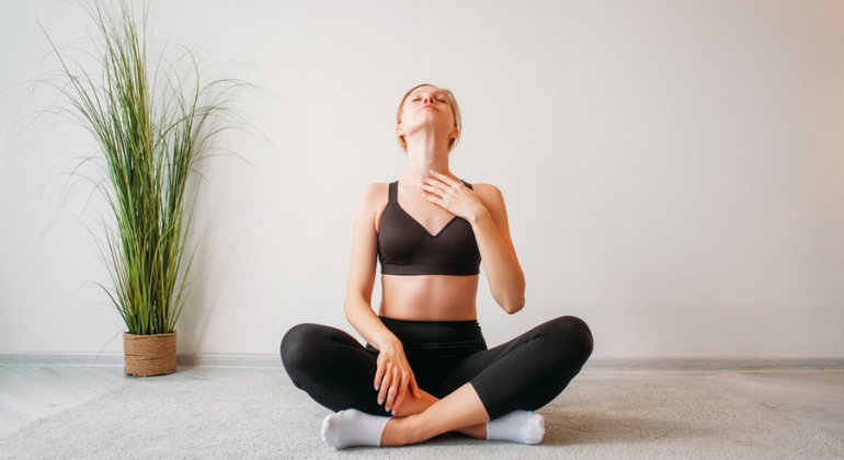 A woman sitting cross-legged on a carpet practicing a neck stretch. She is gently tilting her head back while placing one hand on her chest for support, performing a relaxation exercise in a serene home setting.