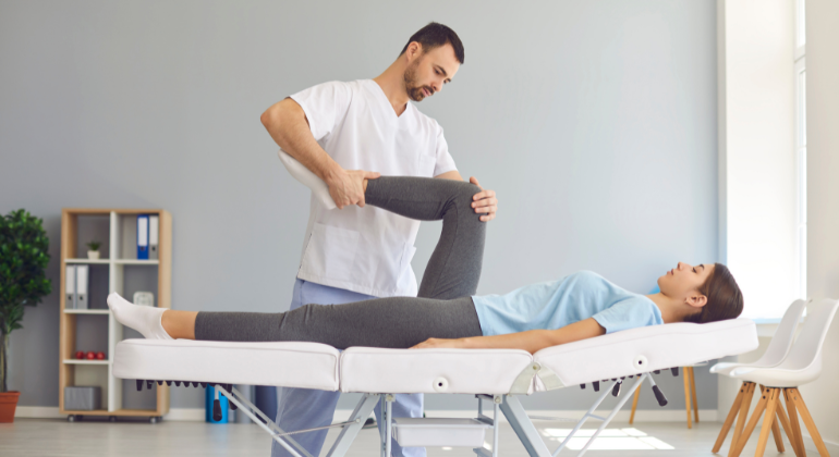 A physiotherapist performing knee rehabilitation exercises on a female patient lying on a treatment table in a clinic. The therapist is gently bending the patient’s leg to assess flexibility and mobility.