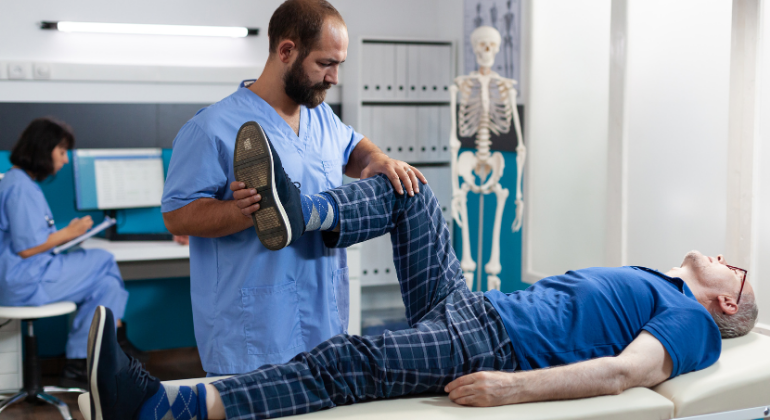 A physiotherapist assisting an elderly patient with a knee mobility exercise during a rehabilitation session in a medical clinic. The patient is lying on a treatment table while the therapist bends his leg to restore movement.