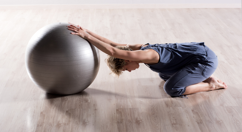 A woman performing a child’s pose stretch using a stability ball. She is kneeling on the floor, extending her arms forward onto the ball in a calm indoor setting.