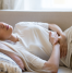 A woman lying on a couch, clutching her abdomen in discomfort, possibly experiencing stomach pain, bloating, or digestive issues.